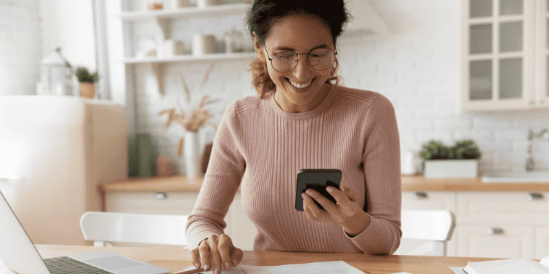 A woman sits at a kitchen table and smiles at her phone.
