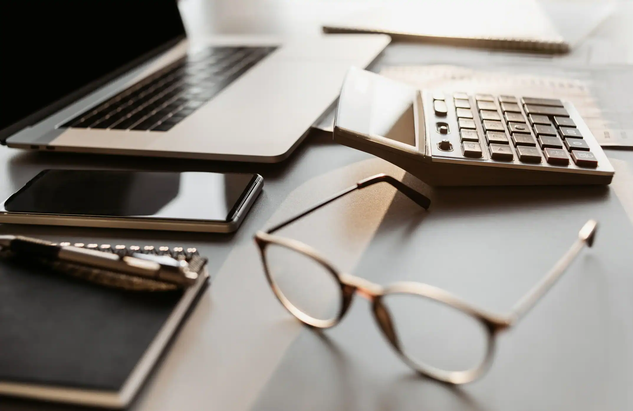 Close-up of a desk with a laptop, calculator, smartphone, notebook, and eyeglasses.