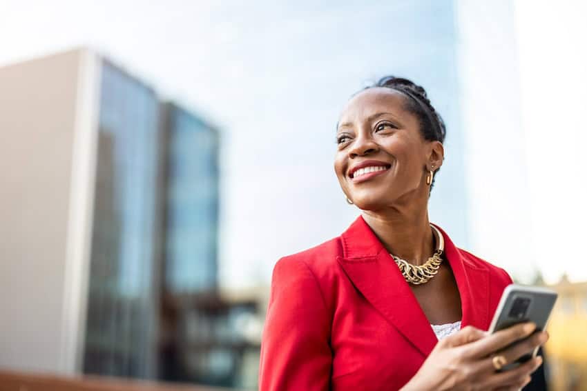 Smiling woman in a red blazer holding a phone outside an office building.