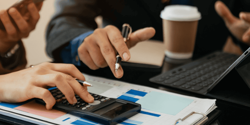 Two people are having coffee and discussing finances. One is using a calculator and the other is writing notes.